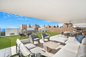 A modern outdoor living area with a sofa, chairs, and a dining table, overlooking a well-maintained lawn and a view of the sea. - Luxury Apartment with Ocean View (Marbella)