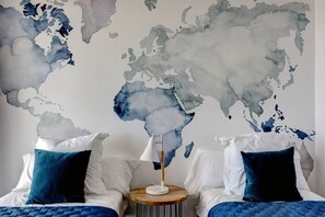 Un dormitorio con un mural de mapa mundial, dos camas con ropa de cama azul y blanca, y una mesita de noche con una lámpara.