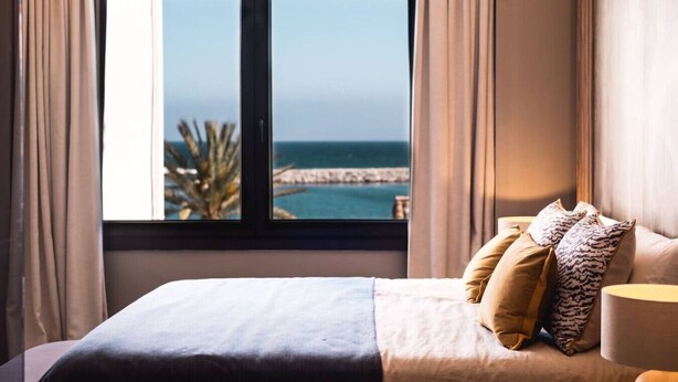 Une chambre à coucher avec un lit, des oreillers, une table de chevet et une vue sur la mer par la fenêtre.