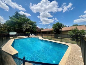 Ein rechteckiger Swimmingpool mit glasklarer blauer Wasseroberfläche, umgeben von einem schwarzen Zaun, und eine überdachte Terrasse mit Gartenmöbeln.