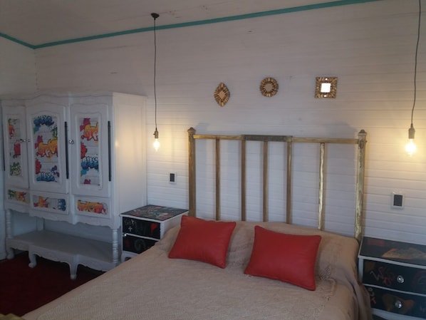 A bedroom with a bed, two red pillows, a white wardrobe with colorful artwork, and a wooden headboard.