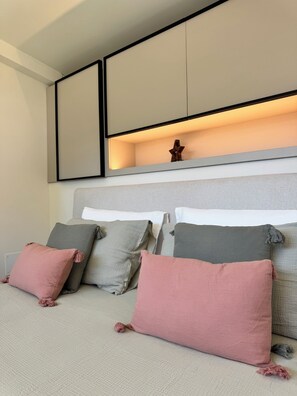 Ein modernes Schlafzimmer mit grauem Kopfteil, mehreren Kissen und einem an der Wand befestigten Regal mit einem dekorativen Gegenstand.