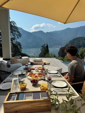 A dining table set outdoors with a scenic mountain and lake view, featuring a variety of food and drinks. - VILLA AQUA LUXURY DESIGN - LAKE COMO VIEW - TREMEZZINA-100 METERS FROM THE SHORE (Tremezzina)