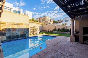 A swimming pool with a tiled deck, a pergola, and a view of modern buildings in the background. - Coastal stay in safest neighbourhood. (Cape Town)