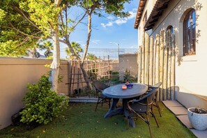 A patio with a round table and chairs, surrounded by a bamboo fence, with a building in the background. - Coastal stay in safest neighbourhood. (Cape Town)