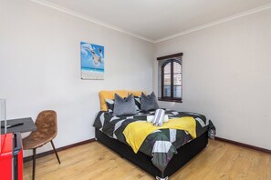 A bedroom with a bed, a desk, a chair, a window, and a painting on the wall. - Coastal stay in safest neighbourhood. (Cape Town)