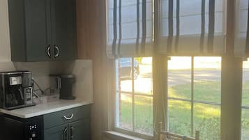 Una cucina con mobili scuri, una macchina da caffè e una sedia in vimini vicino a una finestra con persiane.