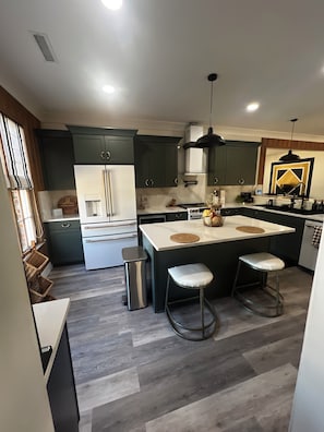 A modern kitchen with dark green cabinets, a white island with stools, and a stainless steel trash can.