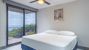 Un dormitorio con cama, ventilador de techo, ventana grande con vista al mar y un cuadro en la pared.