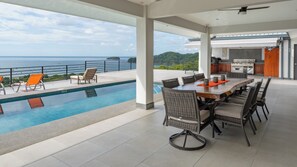 Ein Poolbereich mit Esstisch und Stühlen, Blick auf das Meer und eine Küchenzeile.