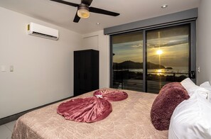 Ein Schlafzimmer mit Bett, Deckenventilator, Klimaanlage, Kleiderschrank und einer großen Schiebeglasdoor mit Blick auf einen Sonnenuntergang.