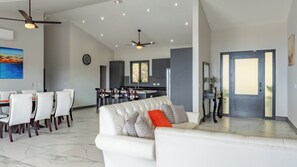 Ein modernes Wohnzimmer mit einem weißen Sofa, einem Essbereich mit Stühlen, einer Küche mit dunklen Schränken und einem großen Fenster.