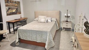 A bedroom with a bed, bedside table, lamp, chair, desk, and a painting on the wall.