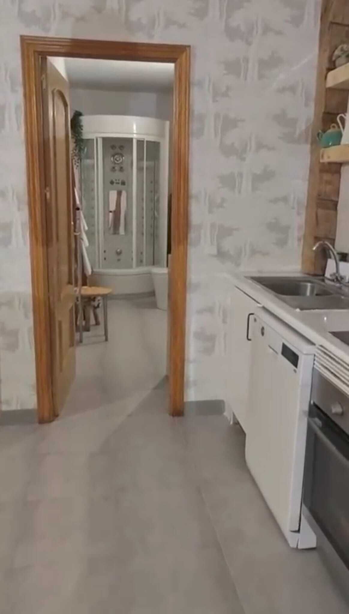 Cocina con armarios blancos, un lavamanos y una estufa. Hay una puerta de madera que da a un baño con ducha e inodoro.