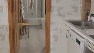 Cocina con armarios blancos, un lavamanos y una estufa. Hay una puerta de madera que da a un baño con ducha e inodoro.