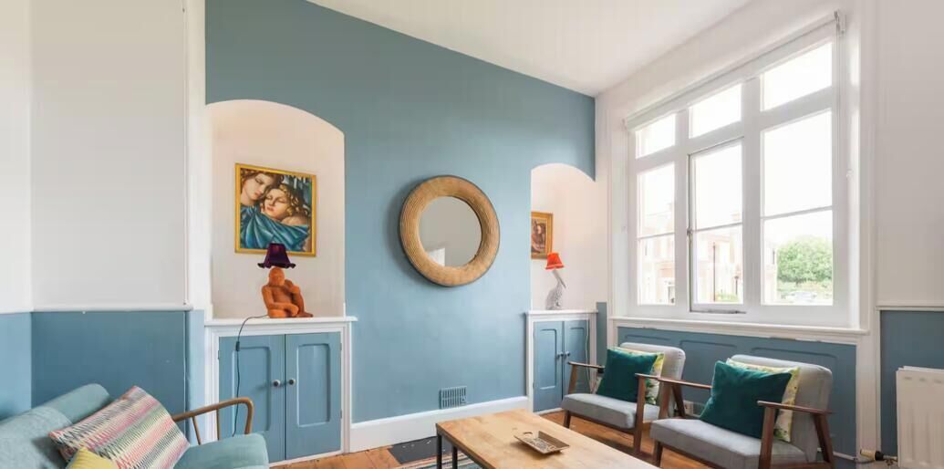 Een kamer met een blauwe wand, een ronde spiegel, een bank, een stoel en een raam met witte kaders.