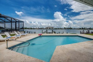 A swimming pool with lounge chairs and a glass-enclosed area, overlooking a body of water with a dock and buildings in the distance. - Villa Fun In The Sun, Lakefront Oasis with Heated Pool & Private Dock........ (Cape Coral)