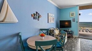 Ein Zimmer mit rundem Tisch, Stühlen, einem Fernseher, Kunstwerken und Blick auf den Strand.