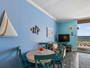 Ein Zimmer mit rundem Tisch, Stühlen, einem Fernseher, Kunstwerken und Blick auf den Strand.