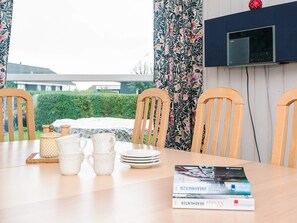A dining table with chairs, a set of white cups and saucers, a stack of plates, and a magazine. A television is mounted on the wall above the table. - 4 Star Holiday Home in Hejls (Hejls)