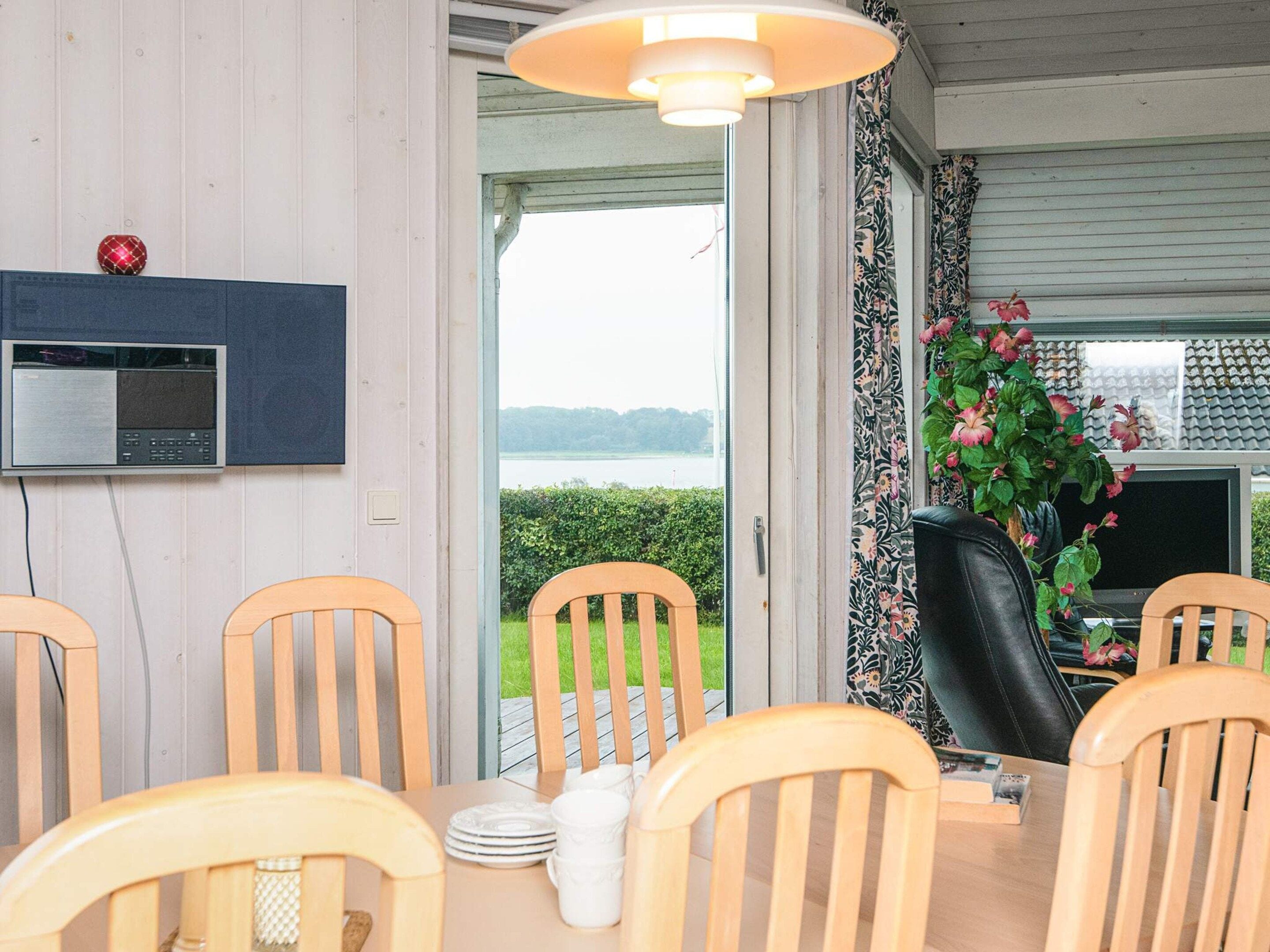 Uma sala de jantar com cadeiras de madeira e uma mesa, uma televisão na parede e vista para um lago através de uma porta aberta.