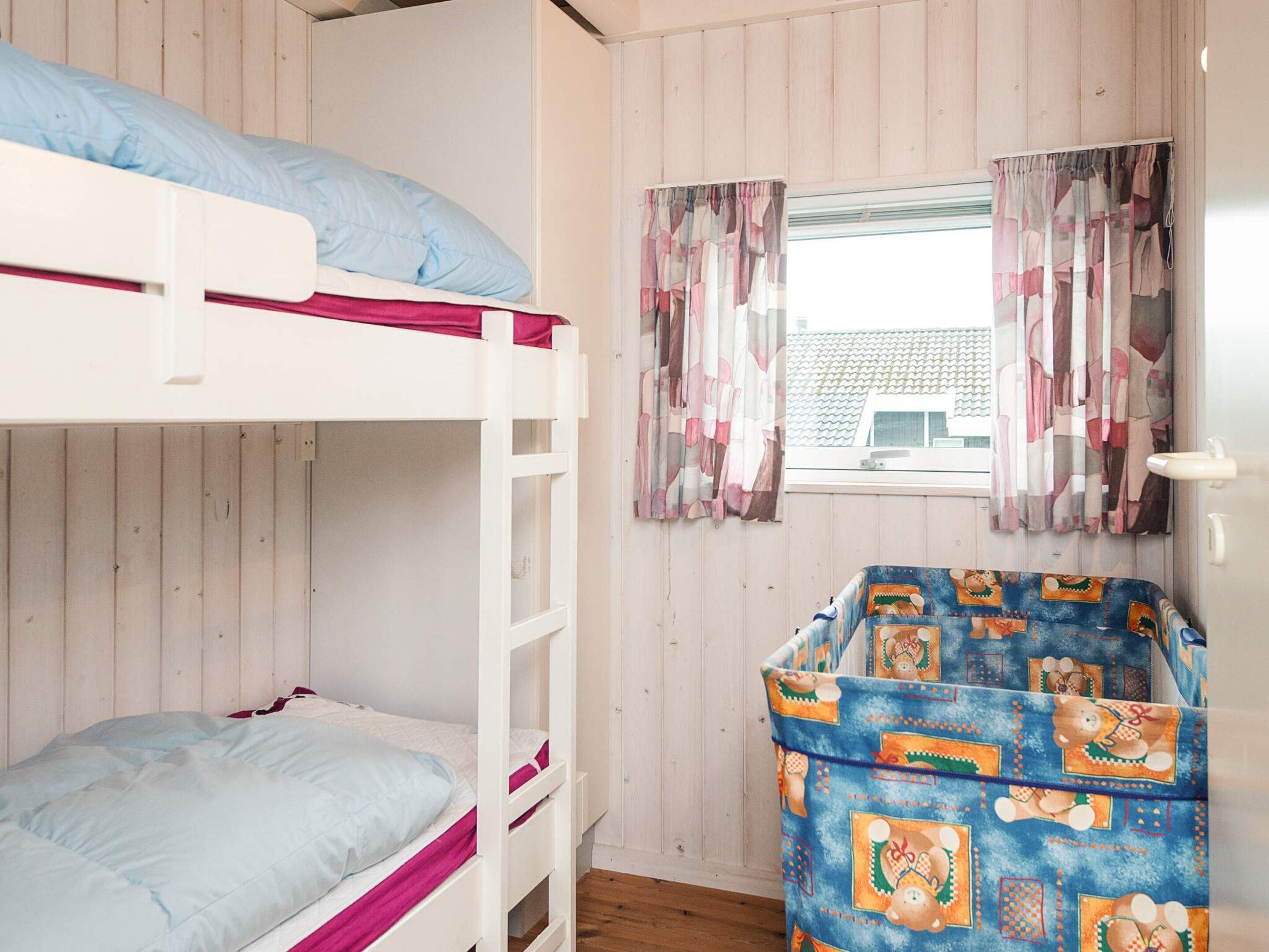 A bunk bed with a blue pillow and a single bed with a pink blanket. There is a window with curtains and a view of a building outside.