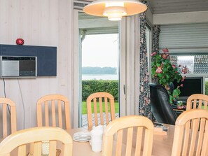 Un coin repas avec des chaises et une table en bois, un téléviseur et une vue sur un lac, visible à travers une porte ouverte.
