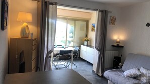 Una habitación con una cama, una mesa y una silla. Se observa una ventana con cortinas, una lámpara y una mesita con un florero.