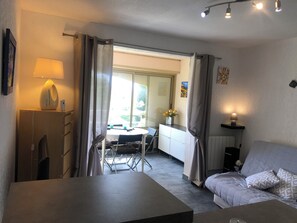 Ein Zimmer mit einem Bett, einem Tisch und einem Stuhl. Es gibt ein Fenster mit Vorhängen, eine Lampe und einen kleinen Tisch mit einem Blumenvasen.