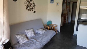 Un salón con sofá, cojines, una mesita redonda y una cocina al fondo.