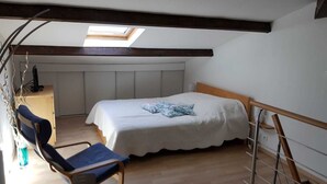 Un dormitorio con techo inclinado, una cama con cubrecama blanco, una silla azul y un escritorio con una computadora.