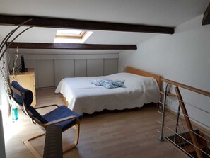 Ein Schlafzimmer mit schrägen Wänden, einem Bett mit weißer Bettdecke, einem blauen Stuhl und einem Schreibtisch mit Computer.