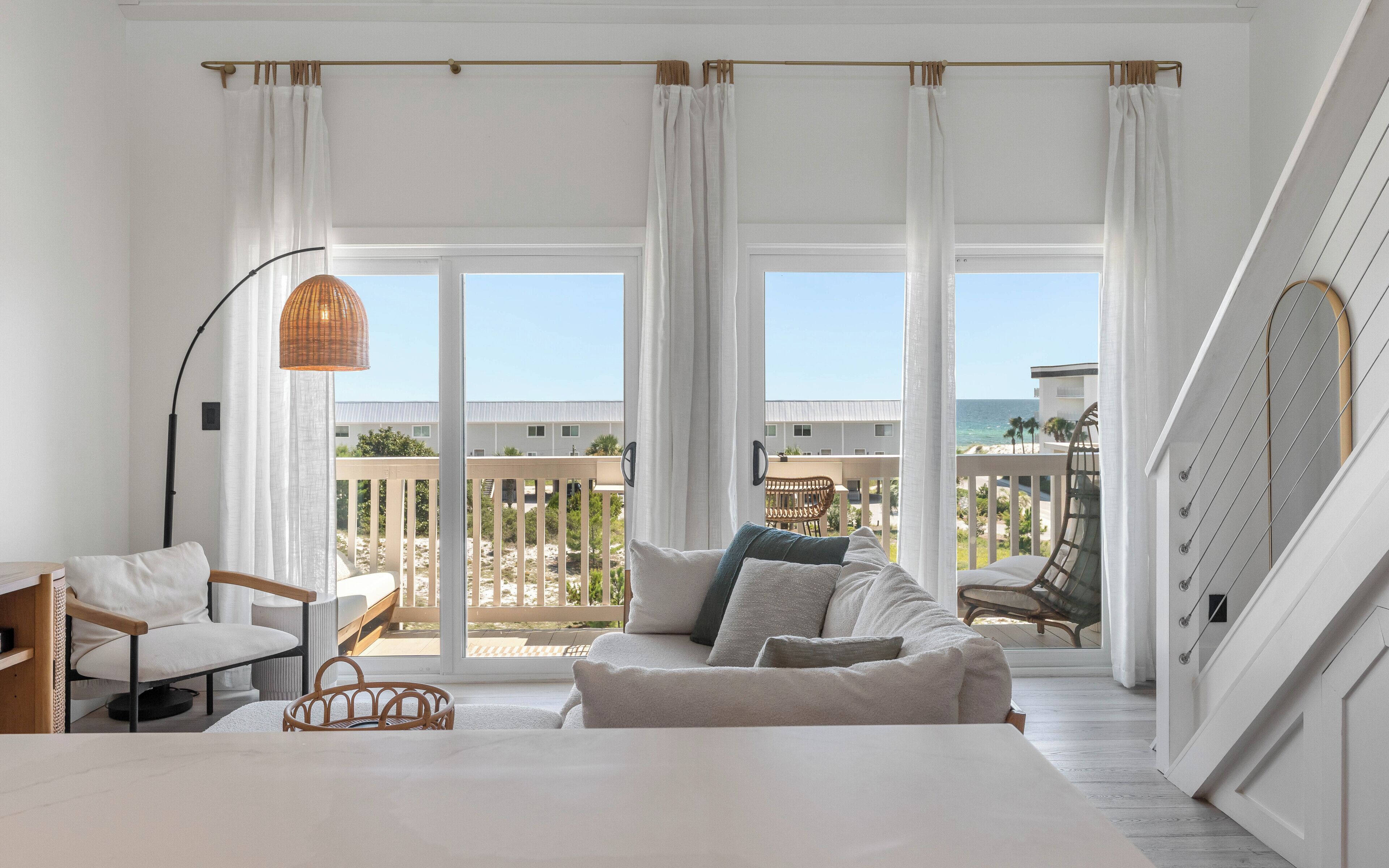 A room with a bed, a seating area with a sofa and chairs, a large window with curtains, and a view of the beach and buildings.