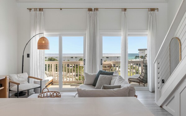 A room with a bed, a seating area with a sofa and chairs, a large window with curtains, and a view of the beach and buildings. - Salt Haus 30A: Pet Friendly Gulf View Loft (Santa Rosa Beach)