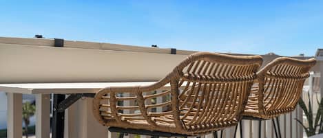 Two wicker chairs with a metal frame, positioned on a balcony with a clear sky in the background.