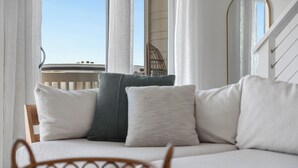 A balcony with a wooden chair, a cushioned sofa with pillows, and a view of the sky and a building. - Salt Haus 30A: Pet Friendly Gulf View Loft (Santa Rosa Beach)