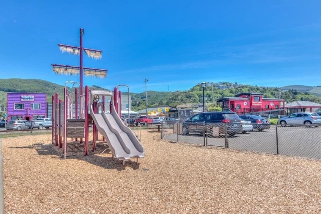 A playground with a slide and climbing structures, surrounded by a parking lot and buildings.