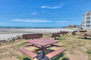 A beachside setting with picnic tables and benches, a building with balconies, and a clear view of the ocean. - Family & Dog friendly Nedonna Beach Home, Hot Tub, BBQ, Short Walk to the Beach (Rockaway Beach)
