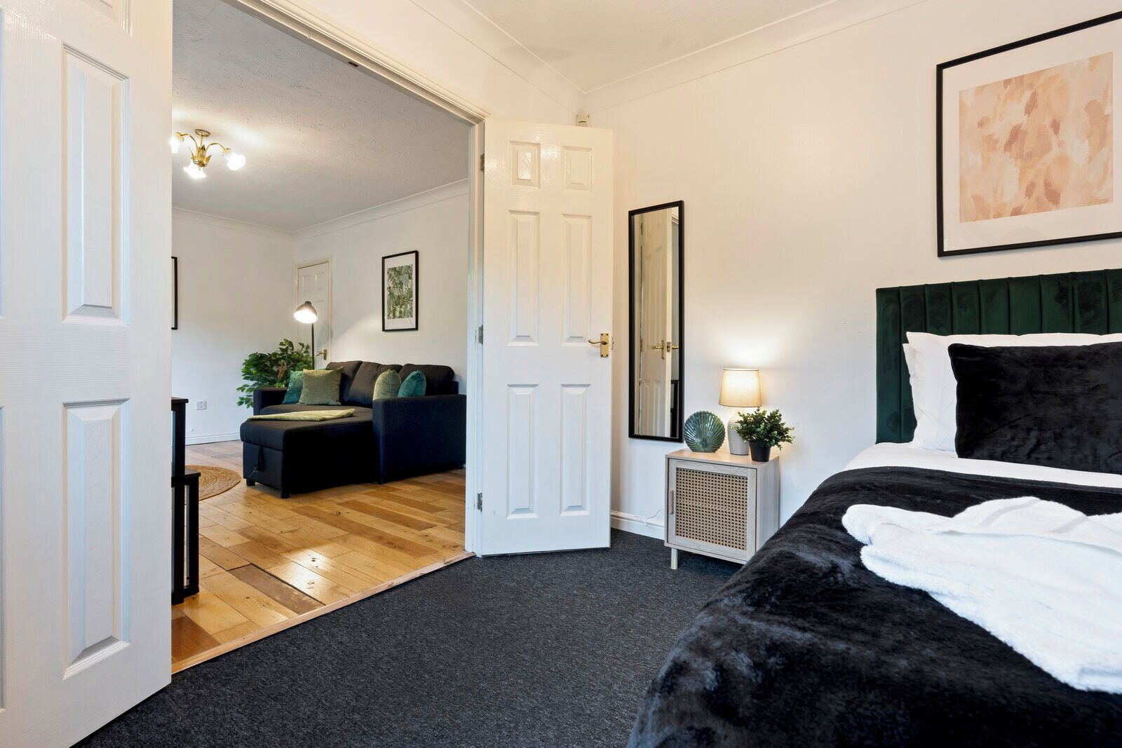 Un dormitorio con cama, mesita de noche con lámpara, espejo y puerta que da a otra habitación.