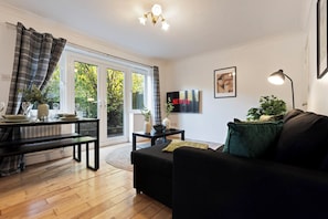 Ein Wohnzimmer mit schwarzem Sofa, einem Esstisch mit Stühlen, einem an der Wand montierten Fernseher und einer Glastür, die in einen Garten führt.