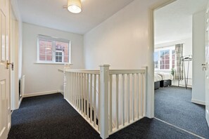 A hallway with a white railing, a window with a brick wall, and a door leading to a bedroom with a bed and a radiator. - Applin House|5BR Contractor Stay|Parking WiFi (Bristol)