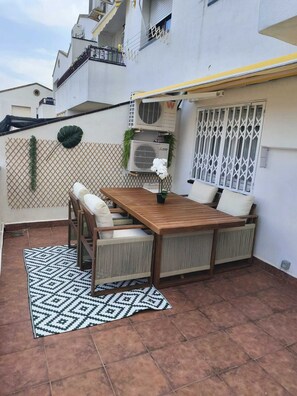 Eine Terrasse im Freien mit einem Holztisch und -stühlen, einem gemusterten Teppich und einer weißen Wand mit Fenster und Klimaanlage.