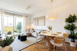 A modern living room with a sofa, dining table, chairs, and a large window. - A light-filled one-bedroom in Cuatro Caminos, where a private sixth-floor balcony looks out over open sky, a quiet place for morning coffee a short walk from the Bernabéu. (MADRID)