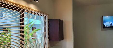 A room with a window with horizontal blinds, a wall-mounted cabinet, and a flat-screen TV on the wall.