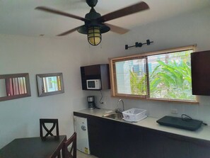 Eine kompakte Küche mit Mikrowelle, Kaffeemaschine und Spüle. Es gibt einen Deckenventilator, ein Fenster mit Jalousien und einen kleinen Essbereich mit einem Stuhl.