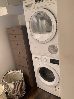 A stacked washer and dryer unit with a control panel, a small trash can, and a cabinet with drawers. - Spacious 170 m² Loft – 3 Bedrooms – Canal St-Martin – Fully Furnished & Designer (Paris)