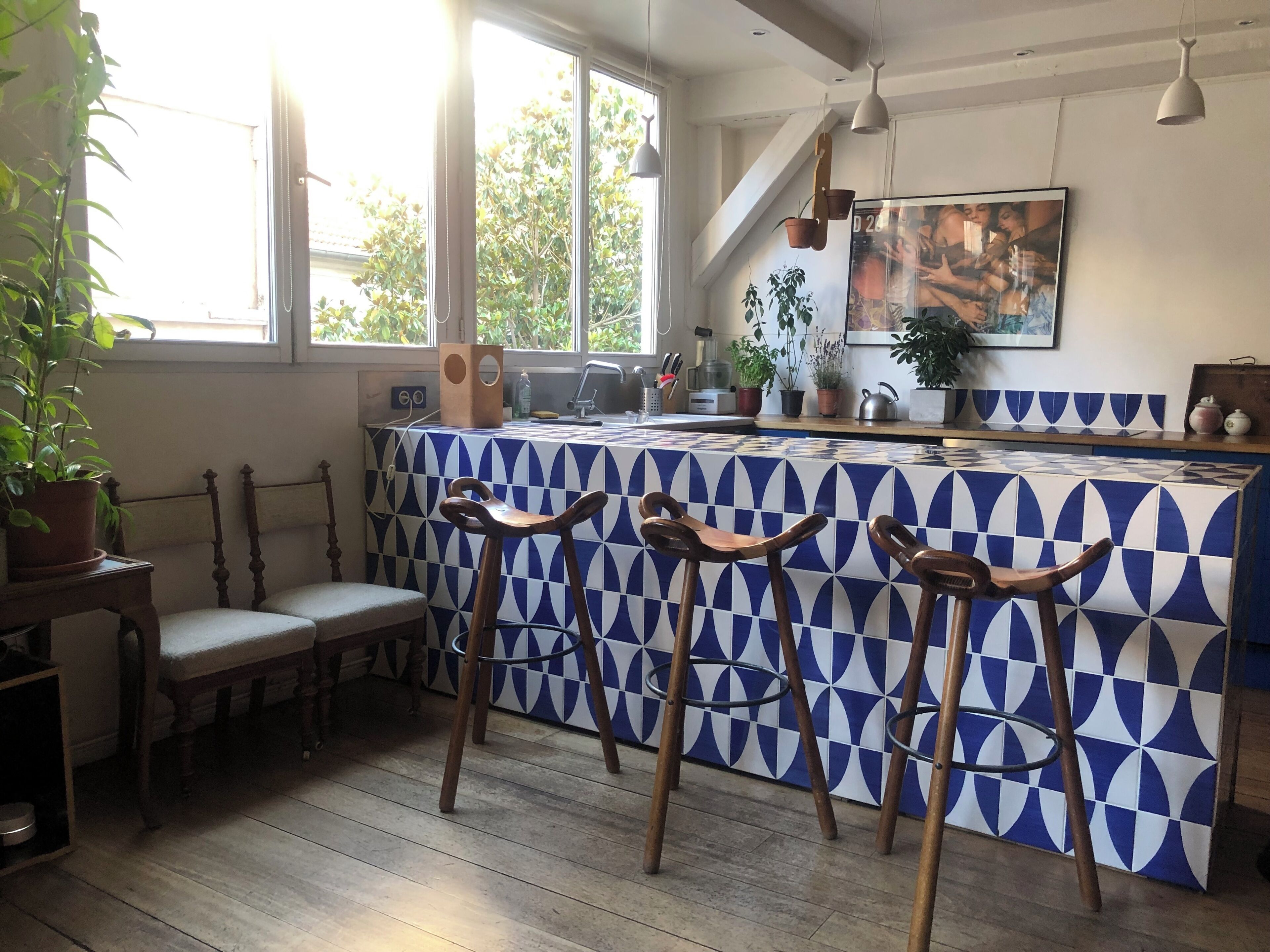 Une cuisine avec un mur recouvert de carreaux bleus et blancs, des tabourets en bois, un comptoir de bar et une fenêtre laissant entrer la lumière naturelle.