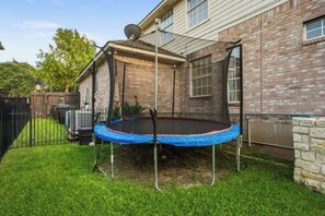 Een achtertuin met een trampoline onder een overdekt gedeelte, een stenen huis en een omheinde tuin.