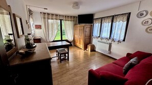 A room with a wooden floor, a red sofa, a dining table with chairs, a television mounted on the wall, and a window with curtains. - Stay at the heart of the slopes in Megève! (Megève)