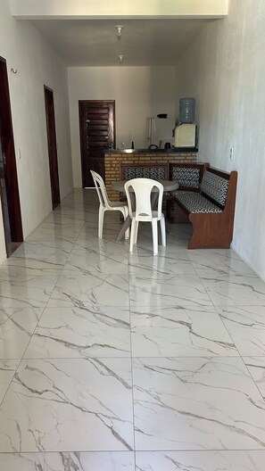 Ein Zimmer mit Marmorboden, einer Holztür, einem weißen Plastikstuhl, einer gemusterten Couch und einer Küchenzeile mit Kühlschrank und Wasserspender.